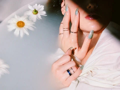 A person's hand elegantly displayed in water, adorned with sparkling handmade fake nails, complemented by fresh daisies and a soft, ethereal glow