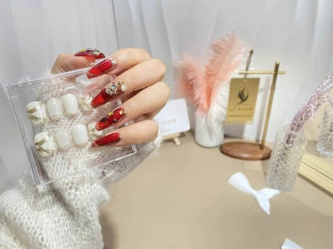 A hand models a striking set of acrylic nails with a bold red and white design, embellished with gold accents and luxurious gemstones, held against an elegant backdrop that includes the LTGlow brand presentation