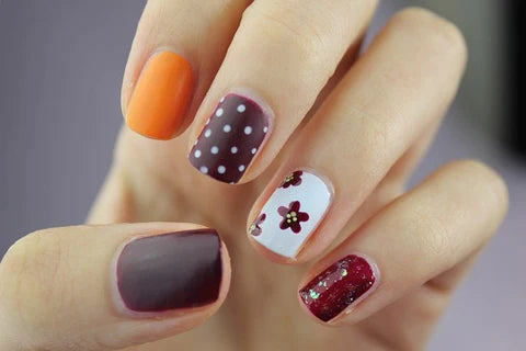 A hand showcasing a variety of handmade press-on nail designs, featuring orange, polka dot, floral, and glitter finishes.