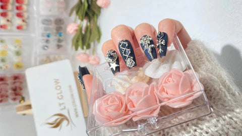 A set of luxurious navy blue and gold press-on nails with intricate floral and gemstone accents, displayed alongside soft pink roses and a LTGlow brand box, emphasizing elegance and craftsmanship.