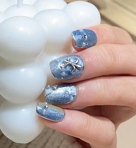 A close-up of a hand showcasing handcrafted press-on nails in a sparkling blue hue, adorned with silver glitter, rhinestones, and a charming bow embellishment, cradling a string of white beads, illustrating the detailed artistry of custom nail design