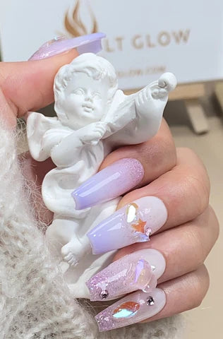 A hand elegantly holding a white cherub statue, showcasing a set of handcraft press-on nails in shades of lavender and pink, adorned with glitter and gemstone accents, with the LT GLOW logo in the background