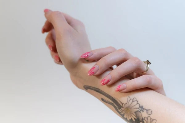 A graceful hand adorned with vibrant pink handmade fake nails, juxtaposed against a prominent black floral tattoo on the forearm. The nails gleam with a light shimmer, and a thin gold ring encircles one finger