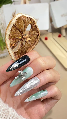 A hand showcasing an array of intricately designed fake nails, with a variety of patterns including black with gemstones, striped silver glitter, and translucent green, all complemented by a dried citrus slice held between the fingers