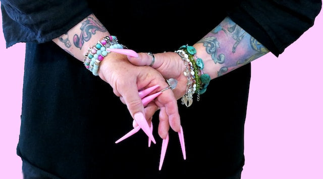 Person showcasing long, pink handmade press-on nails, complemented by an assortment of beaded bracelets and visible tattoo art on their arms