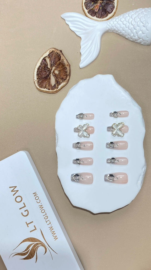 Handmade false nails from LT Glow in a chic coffin shape, showcasing a nude base with 3D butterfly details, glitter, crystal elements, and unique charms